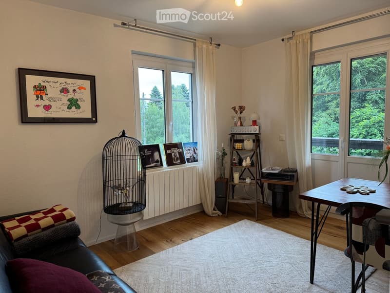 A cozy living room with wooden flooring, large windows, a black leather sofa, a wooden table, a birdcage, a shelf with books and decorative items, and a framed artwork on the wall.