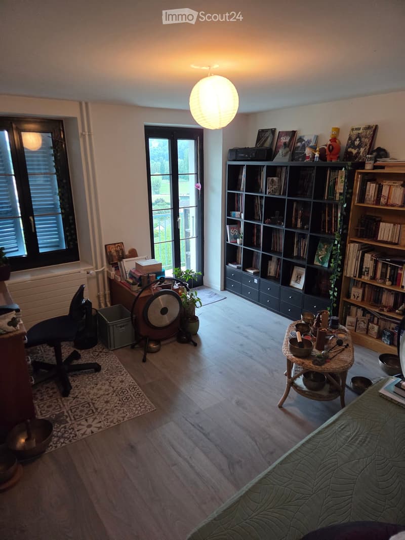 A cozy living room with a green sofa, a wicker side table with bowls, a bookshelf filled with books, a hanging lamp, a chair, and a small table with a plant and decorative items.