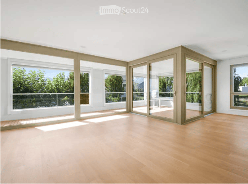 Bright, empty room with large windows and sliding glass doors leading to a balcony, featuring a wooden floor and white walls.