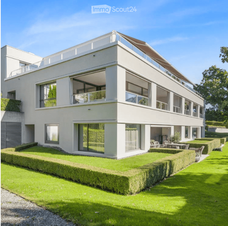 Modern two-story house with white exterior, large windows, and a flat roof. The house features a well-maintained lawn and neatly trimmed hedges in the front yard.