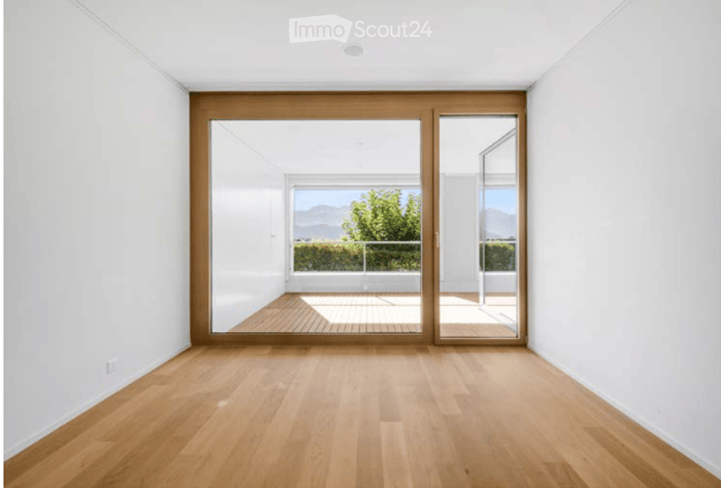 Bright, unfurnished room with light wooden flooring and large glass sliding doors leading to a balcony with a scenic mountain view.