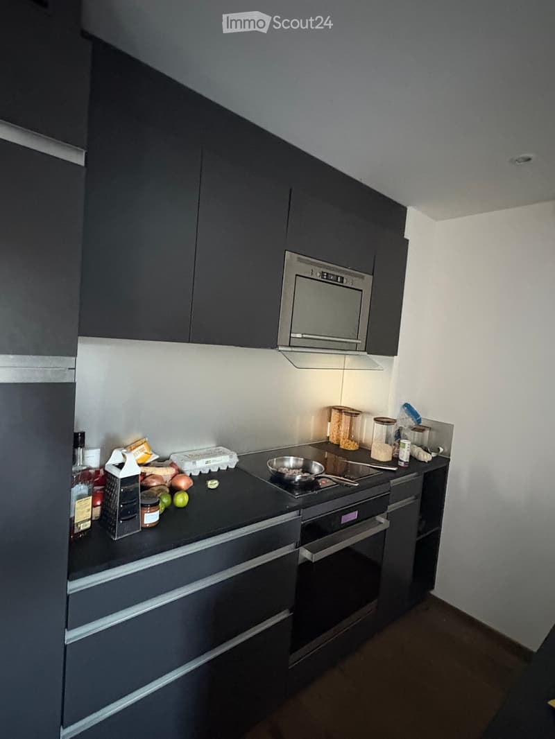 Modern kitchen with dark grey cabinets, stainless steel appliances, and a black countertop. Visible items include a microwave, oven, stove, frying pan, and various food items and kitchen tools.