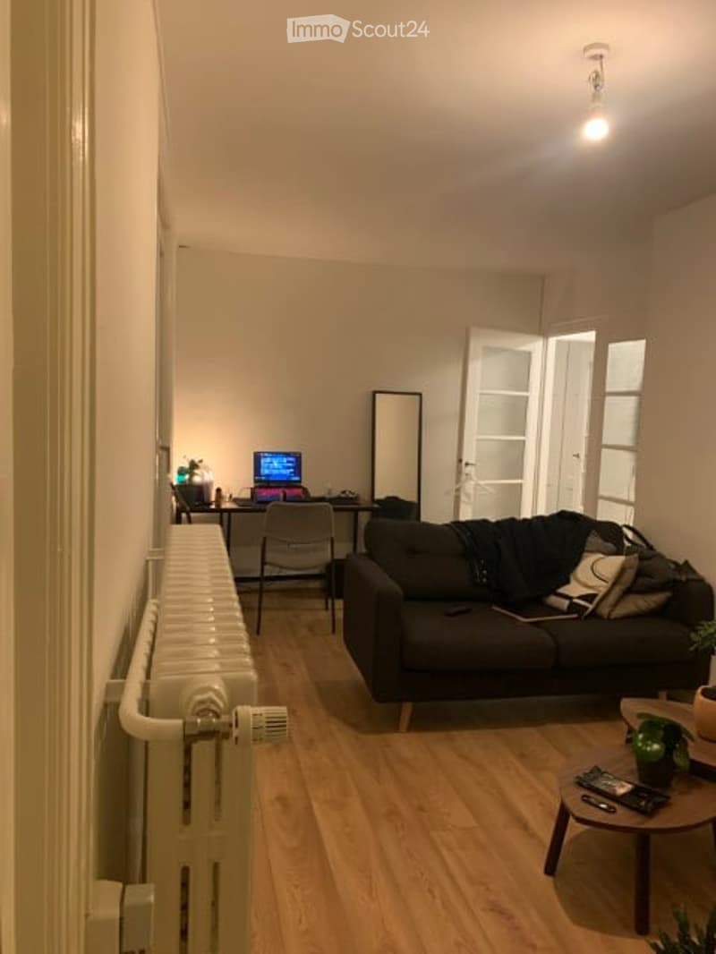 A modern, well-lit living room with wooden flooring, a black sofa, a coffee table, a chair, a laptop on a desk, and a mirror on the wall.