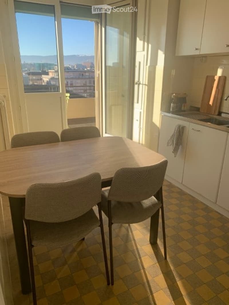 Bright dining area with a round wooden table and four chairs, adjacent to a modern kitchen with white cabinets and a sink. Large windows offer a scenic city view.