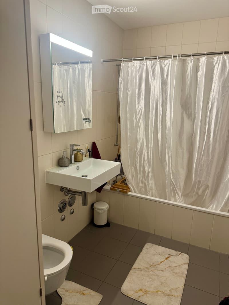 Modern bathroom with white tiles, a sink with a modern faucet, a mirror with built-in lighting, a toilet, a bathtub with a white shower curtain, and marble-style rugs on the floor.