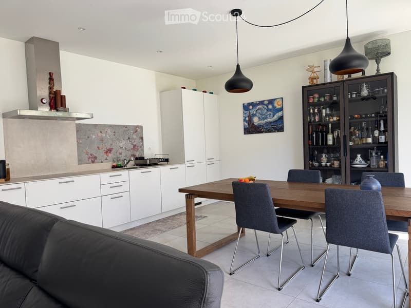 Modern kitchen and dining area with white cabinets, a wooden dining table, grey chairs, a sofa, and pendant lights.