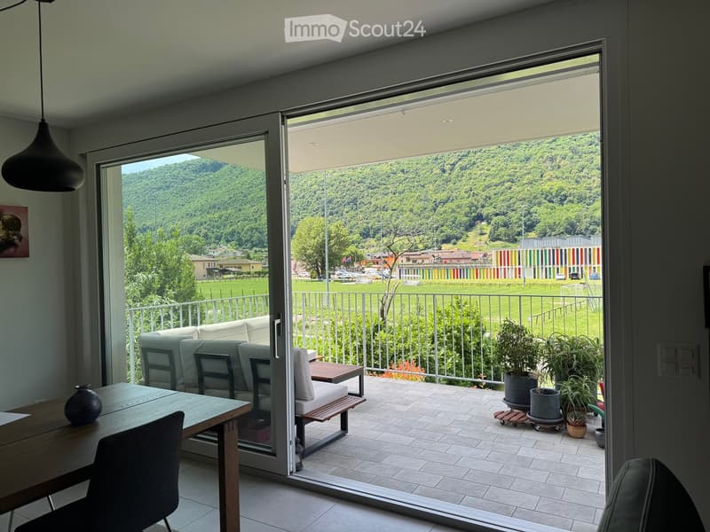 Modern living room with large sliding glass doors opening to a balcony, overlooking a scenic green landscape. The room features a wooden dining table, sofa, chairs, and decorative vases, with potted plants on the balcony.