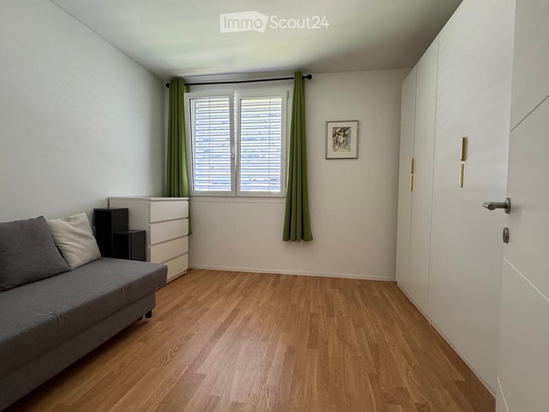 A small, modern living room with a grey sofa, a drawer unit, a side table, and a picture frame on the wall. The room has wooden flooring and a window with green curtains.