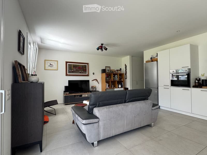 A modern living room with a grey sofa, television, kitchen area with white cabinets, refrigerator, and oven, along with a shelf and a chair.