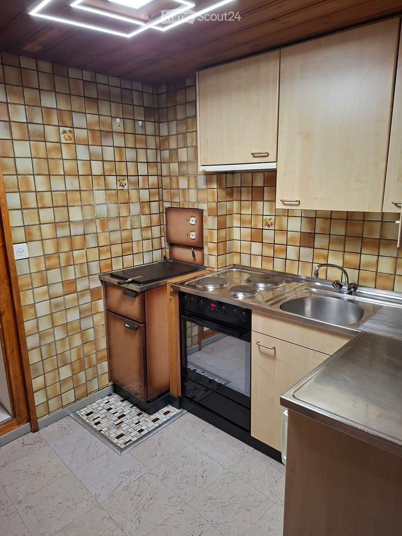 Compact kitchen with light wooden cabinets, a stove, sink, and oven. The walls are tiled with a warm, earthy pattern, and the floor is tiled with a light, speckled design.