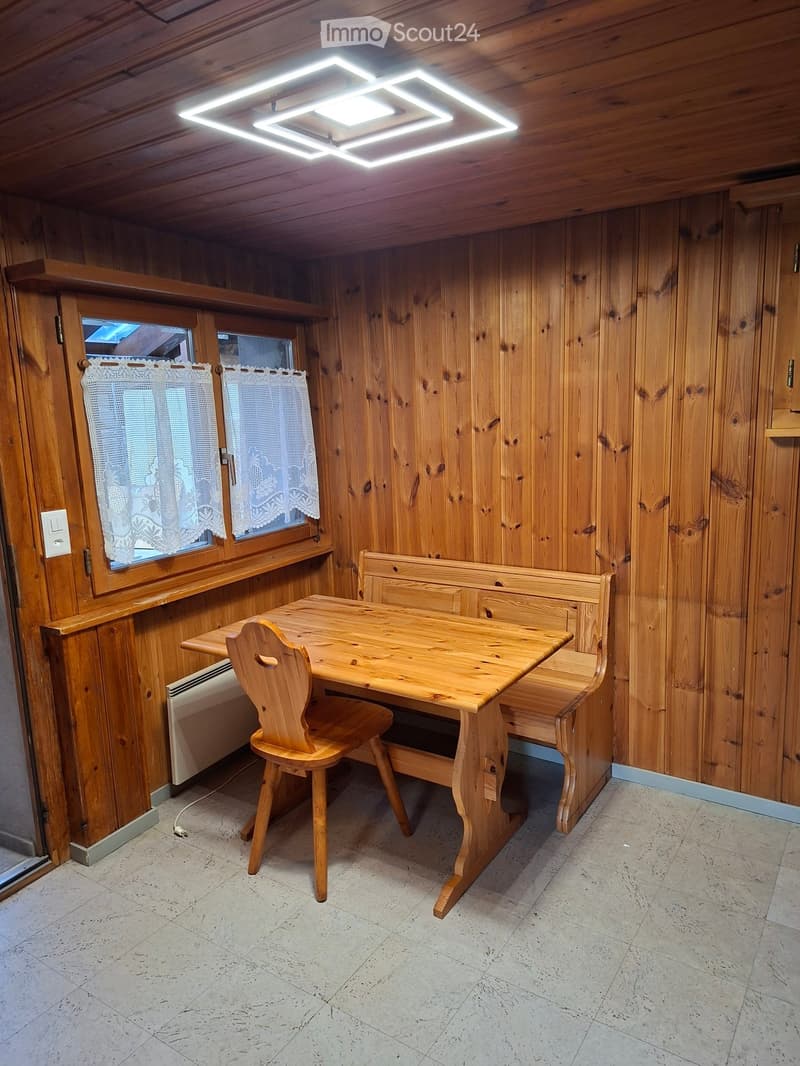Cozy wooden-paneled room with a small dining area featuring a wooden table, a chair, and a built-in bench. The room has wooden walls, a window with lace curtains, and a modern LED ceiling light.