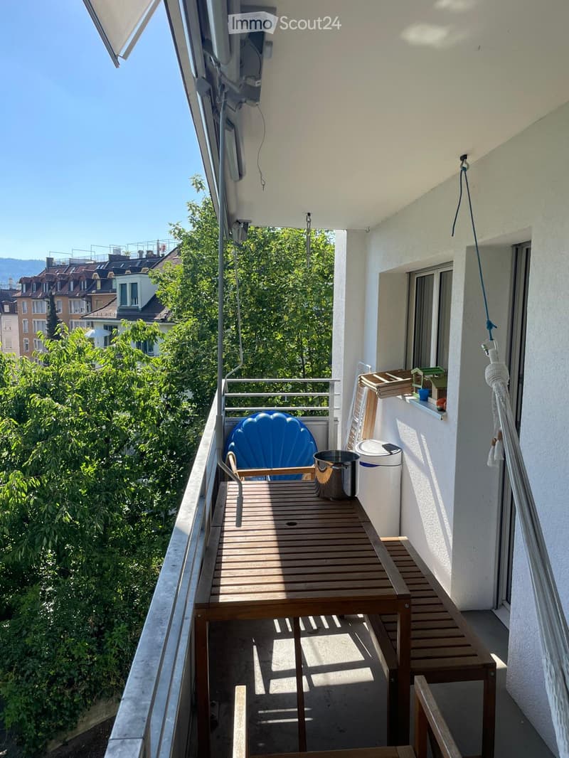 Balcony with wooden table, blue chair, potted plant, and railing overlooking greenery.