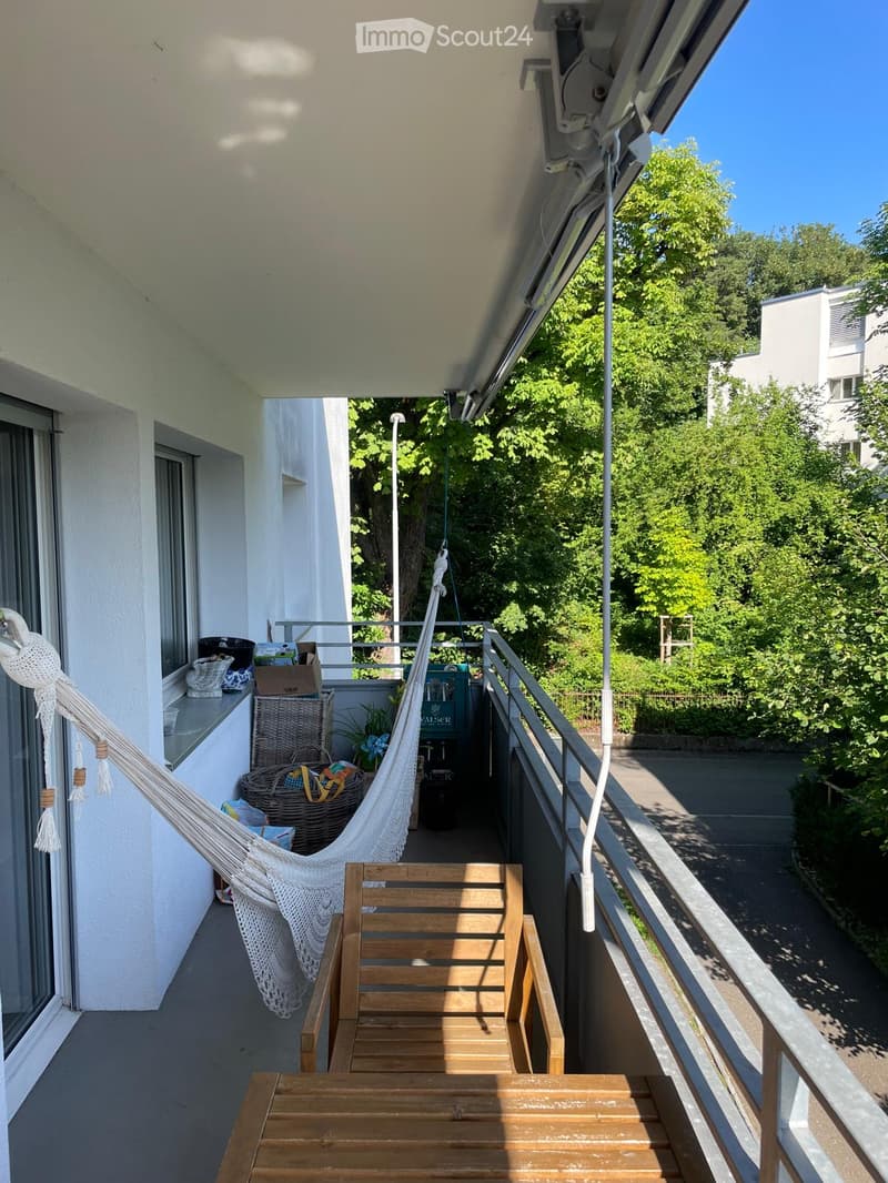 Balcony with wooden chair, table, and hammock, overlooking a green area with trees.