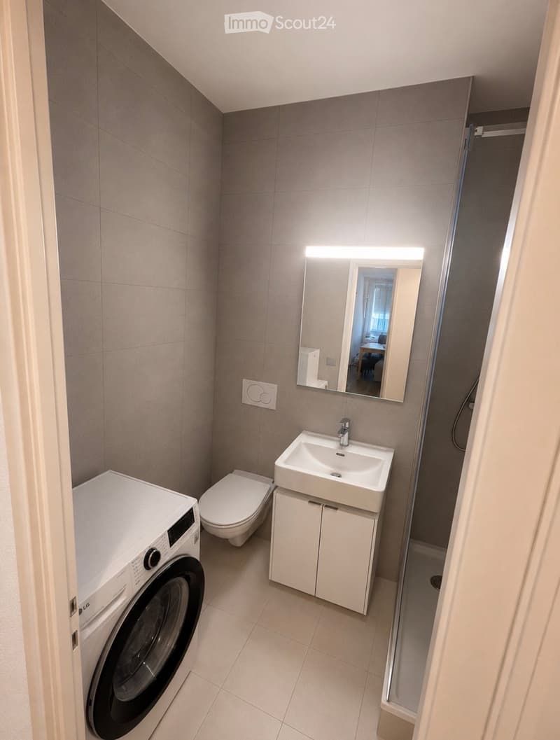 Modern bathroom with a white sink, mirror, toilet, and washing machine. The space features tiled walls and a sleek, contemporary design.