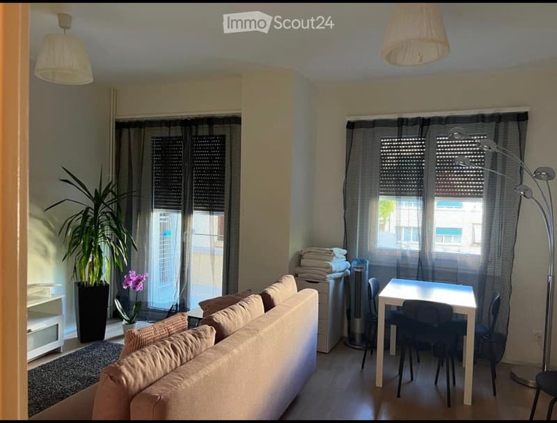 A well-lit living room with a beige sofa, a small dining table with chairs, a floor lamp, and a potted plant near the window.