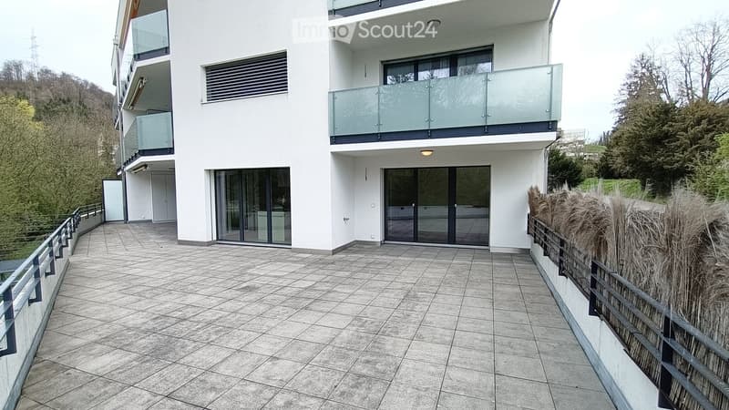 Modern two-story building with white facade, large windows, and balconies. The ground floor features a tiled patio area with a metal railing and a small garden with tall grasses.
