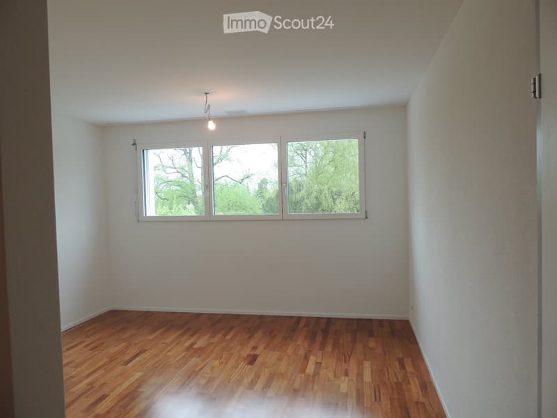 Empty room with wooden flooring, white walls, and large windows showing greenery outside.