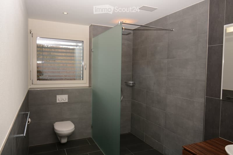 Modern bathroom with dark grey tiles, a white toilet, a glass-enclosed shower, a wooden bench, and a window with blinds.