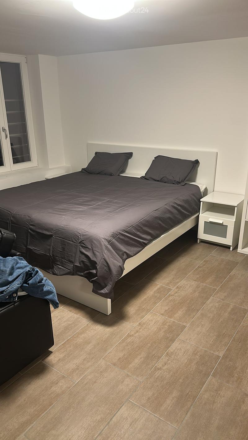 Modern bedroom with a large bed, dark bedding, nightstand, and tiled flooring.