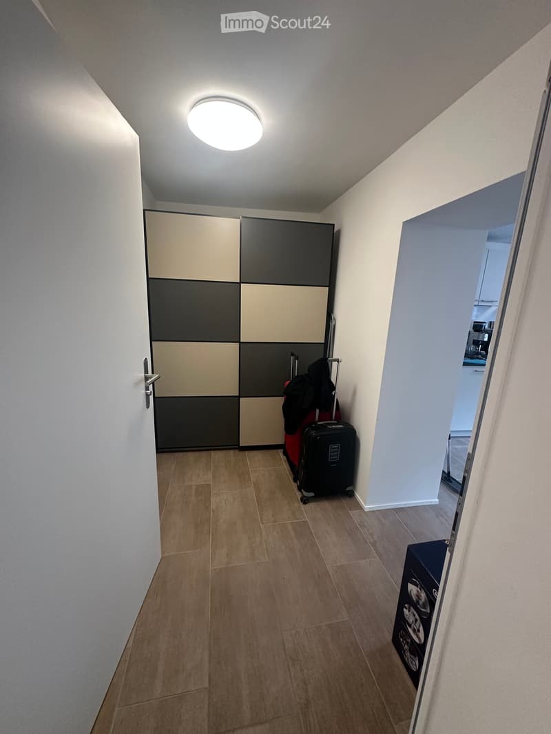 A narrow hallway with tiled flooring, white walls, and a round ceiling light. A large wardrobe with a geometric design is visible on the left, and an open doorway leads to another room on the right.