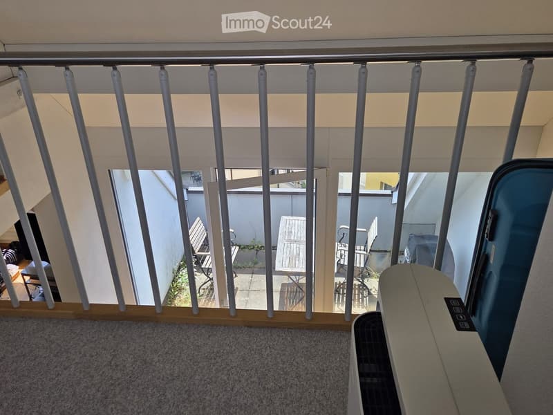 Balcony with grey metal railings, carpeted floor, and a view of a small outdoor seating area with chairs and a table.
