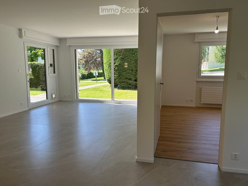 Empty room with tiled floor, large sliding glass doors leading to a green outdoor area, and a wooden floor area visible through an open doorway.