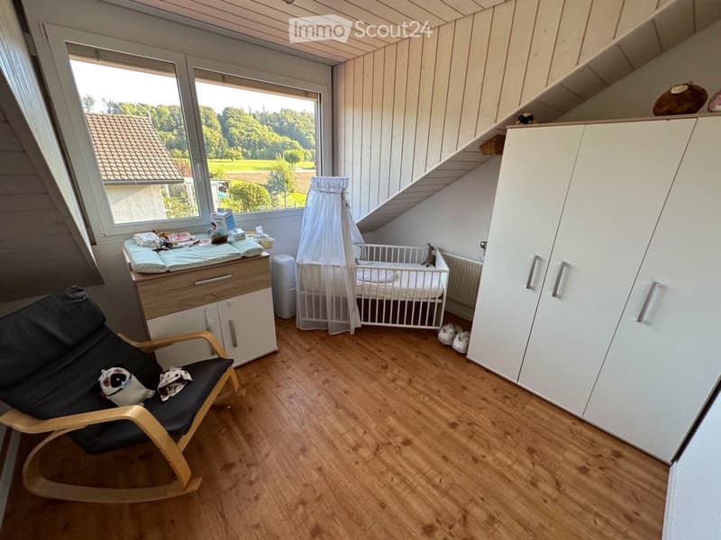 Brightly lit bedroom with wooden flooring, large windows, a baby crib with a canopy, a changing table, a rocking chair, and a white wardrobe.