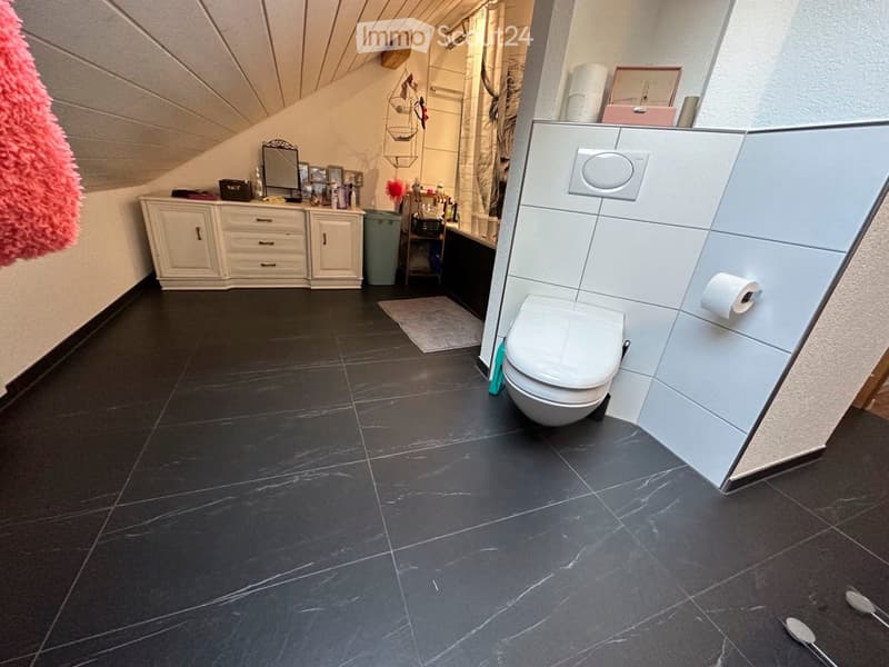 Modern bathroom with dark tiled floor, white toilet, vanity cabinet, mirror, and various toiletries. Features a sleek design with a slanted ceiling and storage space.