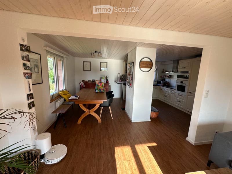 Bright living and dining area with wooden flooring, a large wooden dining table with chairs, a bench, a kitchen area with cabinets, appliances, and a small plant in the corner.