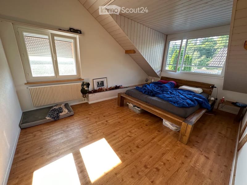 Cozy attic bedroom with wooden flooring, slanted ceilings, and large windows. Features a wooden bed with blue bedding, a radiator, a picture frame, and a small dog bed.