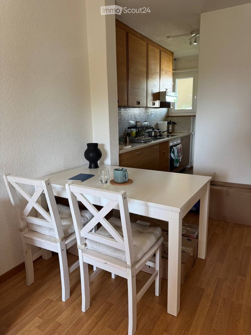 A modern kitchen with wooden cabinets, a white dining table with two chairs, a vase, a cup, and a glass on the table. The kitchen features a stove, sink, and various appliances.
