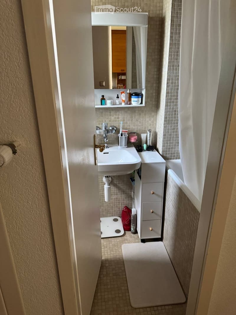 Compact bathroom with a white sink, mirror, small cabinet, and a toilet. The space includes a bathroom scale, a rug, and a shower curtain.