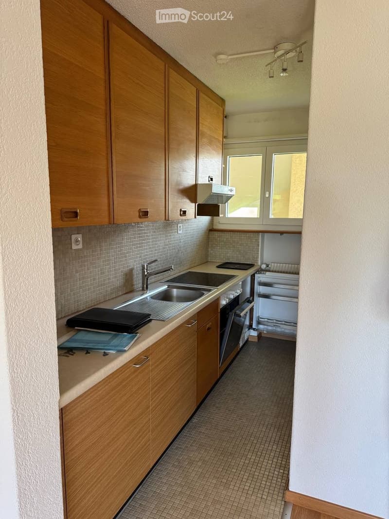 The kitchen features wooden cabinets, a stainless steel sink, a built-in oven, and a refrigerator. The countertop has a few items, including a black laptop and a blue folder.