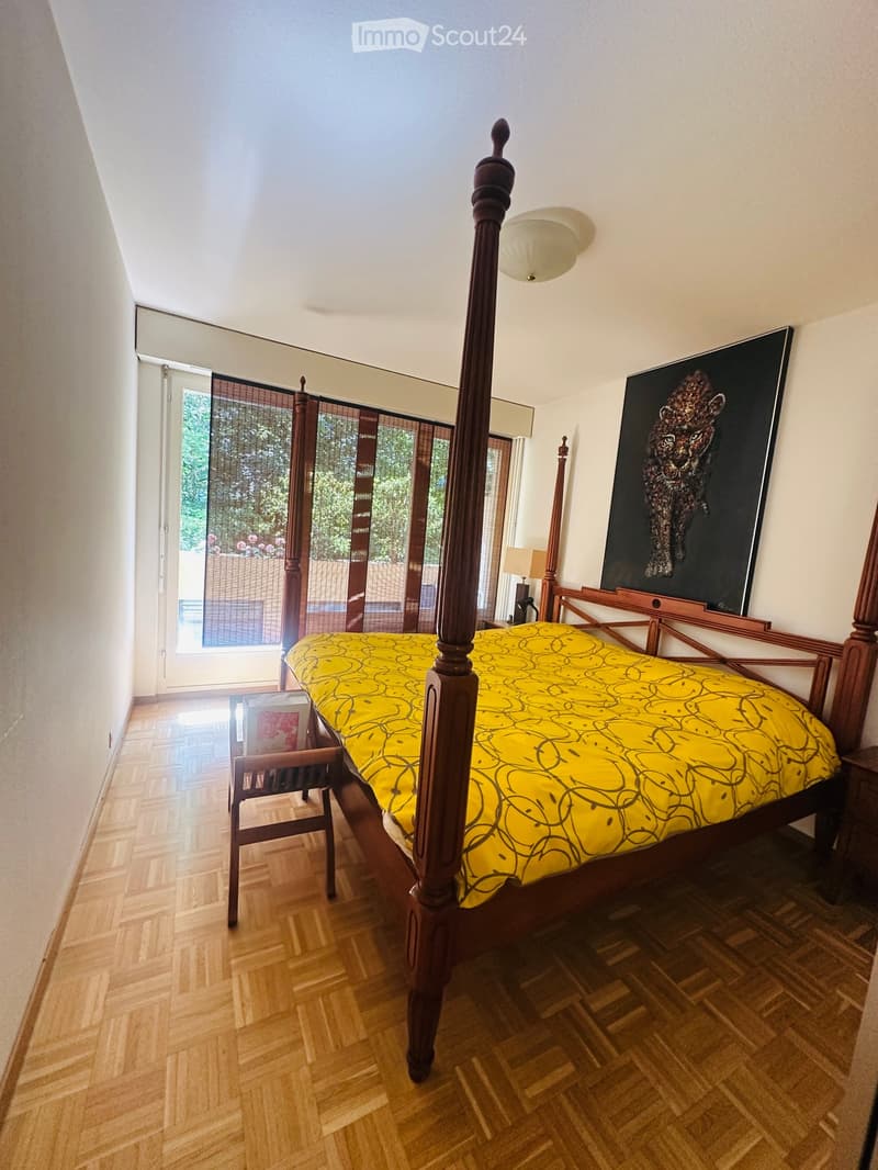 Bright bedroom with wooden bed, yellow bedspread, wooden chair, nightstand, and large painting on the wall. Large window with blinds offers a view of greenery outside.