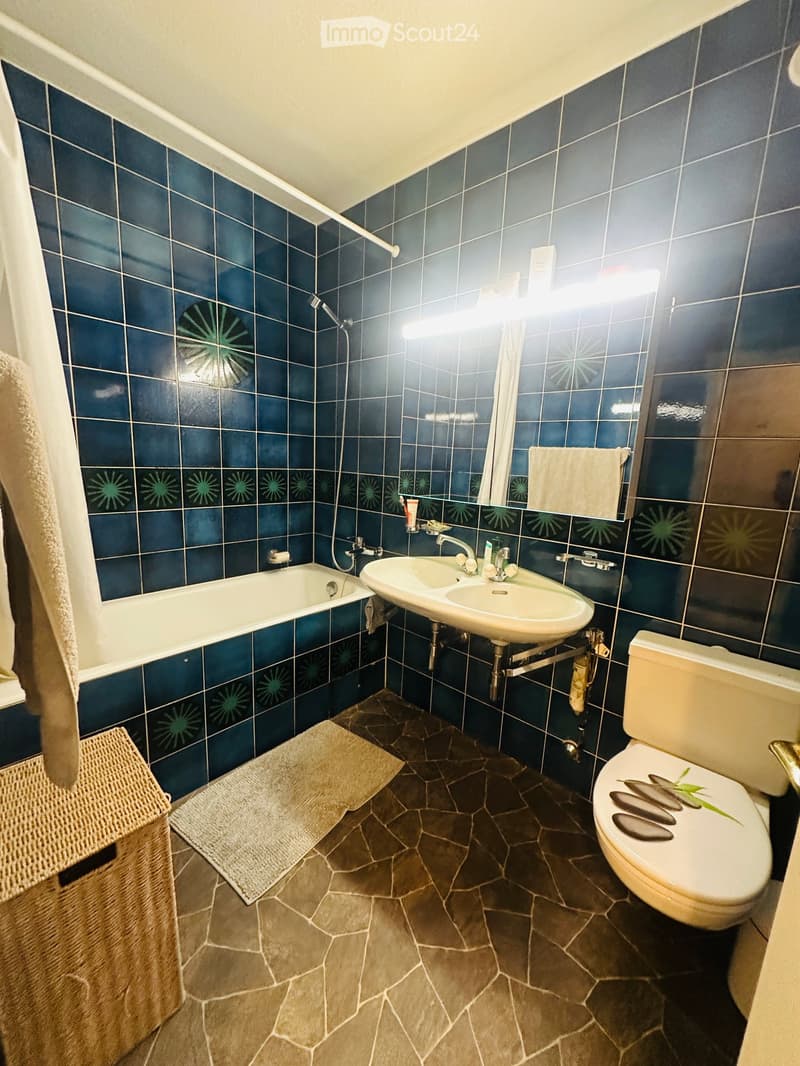 Bathroom with dark blue tiled walls, a bathtub, a sink, a toilet, a mirror, and a towel on the side. The floor is made of dark stone tiles.