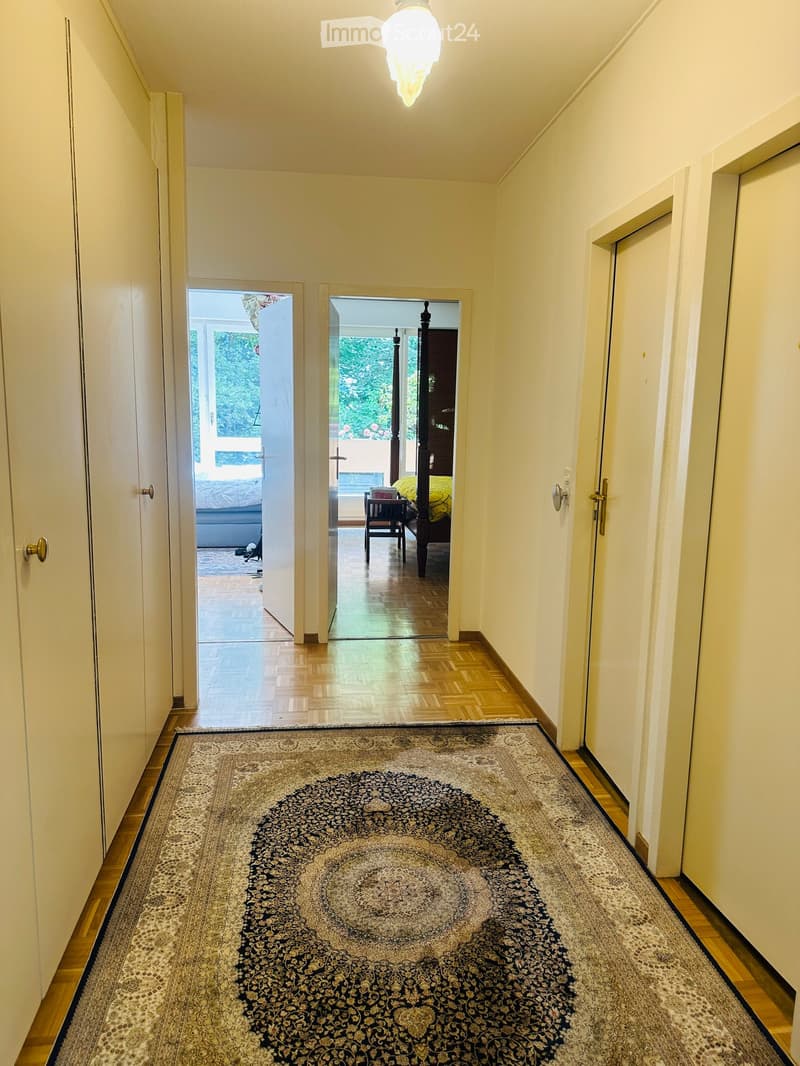 A well-lit hallway with wooden flooring, cream-colored walls, and a large ornate rug in the center. Doors lead to rooms with visible furniture, including a bed and a nightstand.