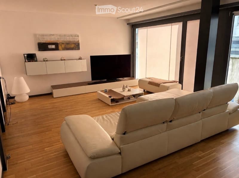 Modern living room with light-colored furniture, including a large sectional sofa, coffee table, ottoman, and a TV stand. The room features a sleek wall-mounted cabinet and a contemporary floor lamp.