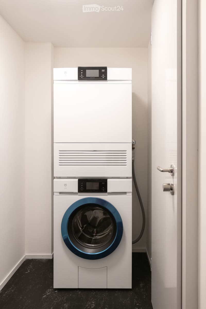 A small, modern laundry room with a white washing machine and dryer stacked vertically, featuring a sleek design and digital control panels.