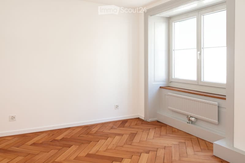 Empty room with white walls, wooden herringbone flooring, large windows, radiator, and electrical outlets.