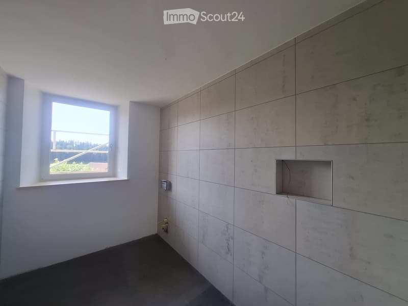 Empty room with light grey tiled walls and a dark grey tiled floor. A single window with a white frame is visible on one wall, and there is a small built-in shelf on another wall.
