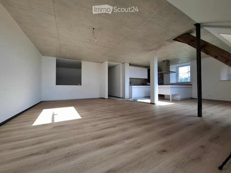 Open, unfurnished room with wooden flooring, white walls, and exposed wooden beams on the ceiling. The room features a large window allowing natural light to enter, and a small kitchen area is visible in the background.