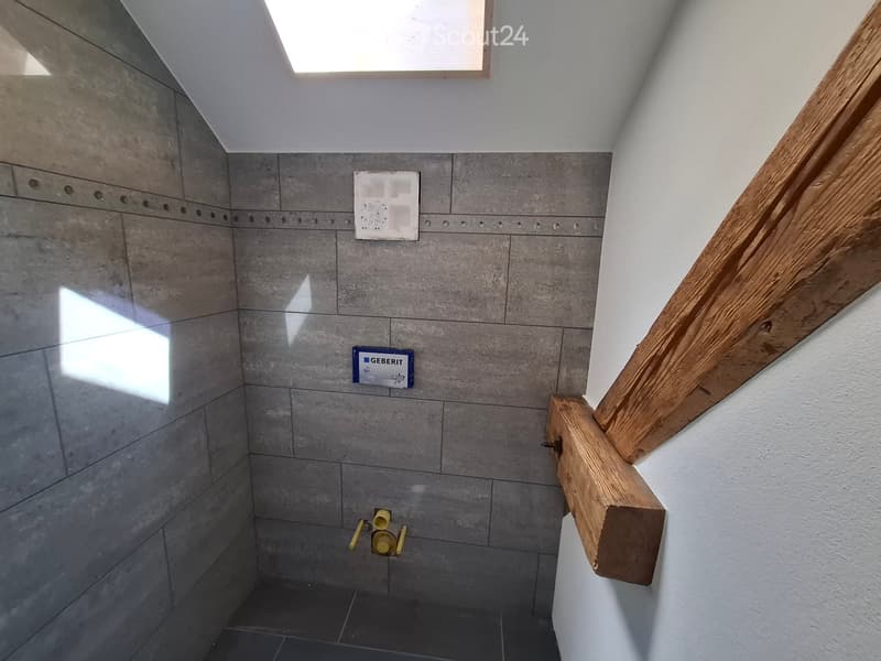 Empty room with grey tiled walls, dark floor, wooden beam, and small window at the top.