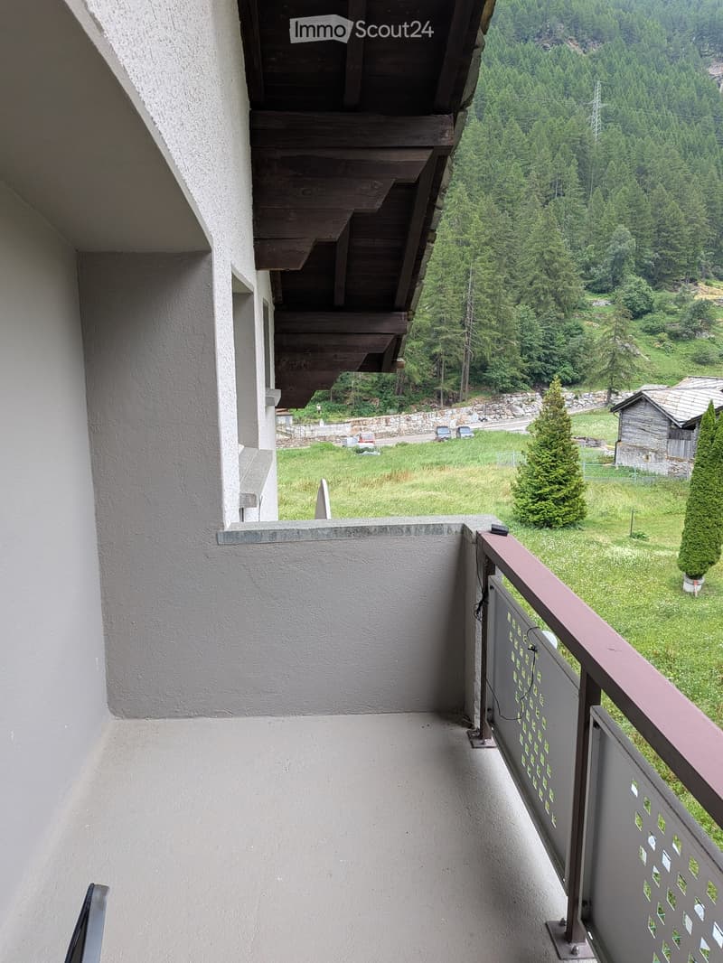 Balcony with wooden staircase leading up, metal railing with decorative pattern, overlooking a green landscape with trees and a small wooden structure.