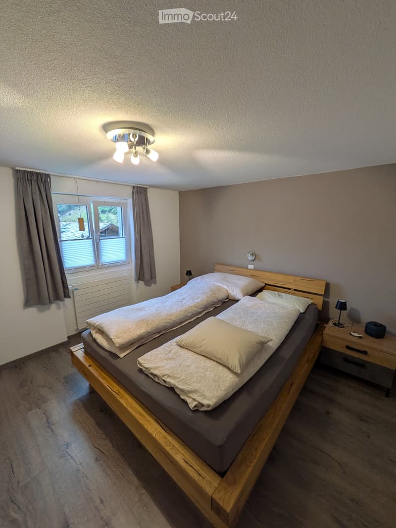 Cozy bedroom with a double bed, two pillows, and a nightstand with a lamp. The room features wooden flooring and a window with curtains.