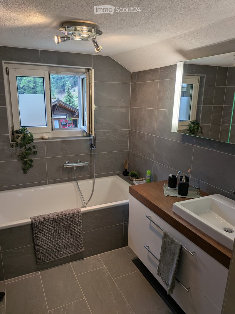 Modern bathroom with a bathtub, sink, mirror, and wooden countertop. Features grey tiles, a window with a view, and decorative plants.