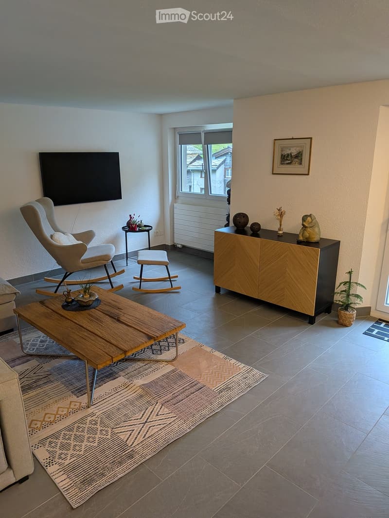 Modern living room with a grey sofa, wooden coffee table, armchair, small table, and decorative items on a cabinet.