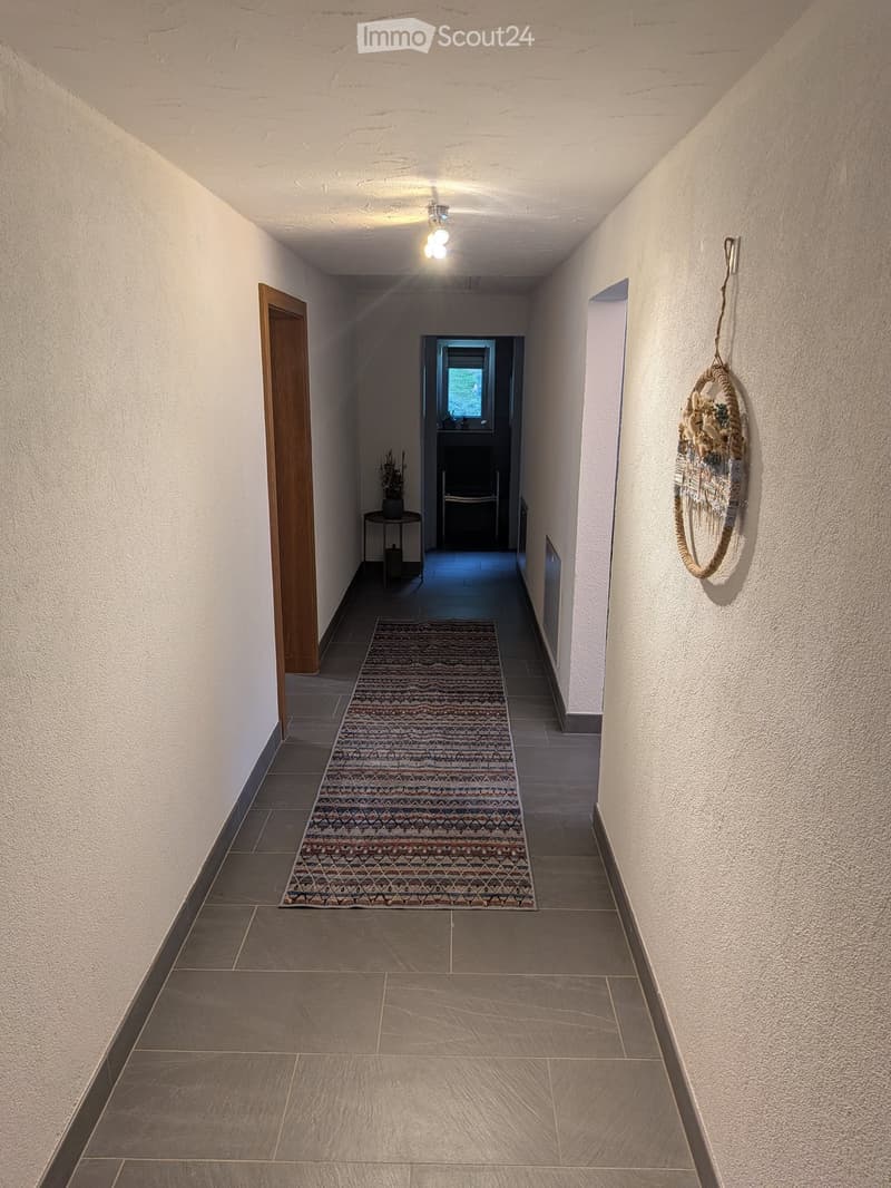 A narrow hallway with tiled flooring, white walls, and a patterned runner rug. A small table with a plant sits near a doorway at the end, and a decorative hanging ornament is visible on the right wall.
