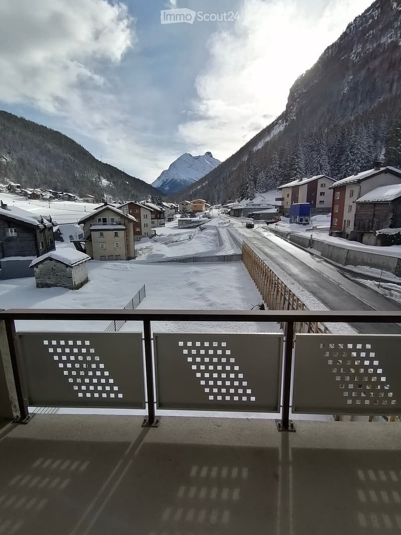 Balcony overlooking a snow-covered village with houses, a road, and distant mountains under a partly cloudy sky.