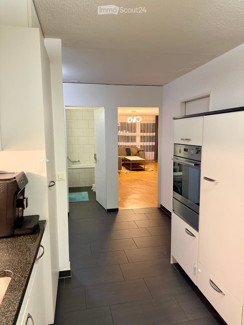 A modern hallway with tiled flooring, white walls, and a view into a kitchen and living area. The kitchen features white cabinets, a coffee machine, and a built-in oven. The living area has a sofa, coffee table, and a chandelier.