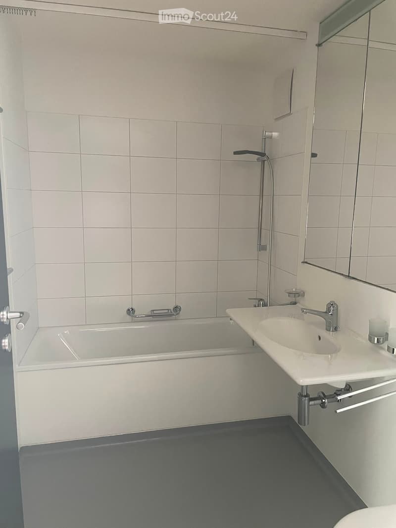 Modern bathroom with a bathtub, sink, and mirror. The walls are tiled in light gray, and the floor is dark gray.
